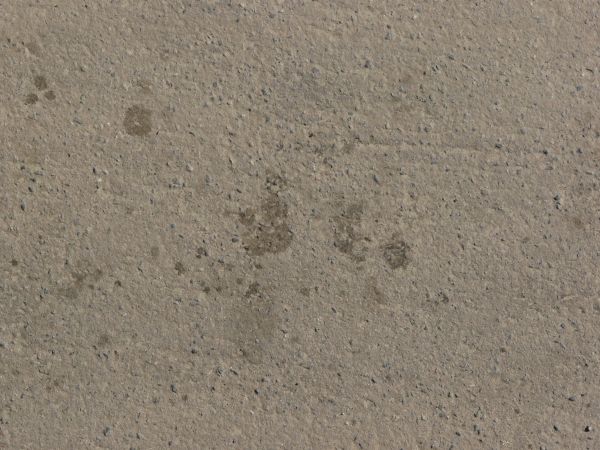Asphalt texture with a coarse tan surface embedded with black stones, and with very little scratching. Several damp, discolored spots are visible near the center.